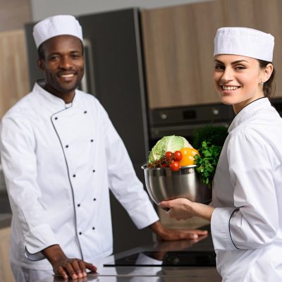 multicultural chefs holding vegetables and looking at camera at restaurant kitchen